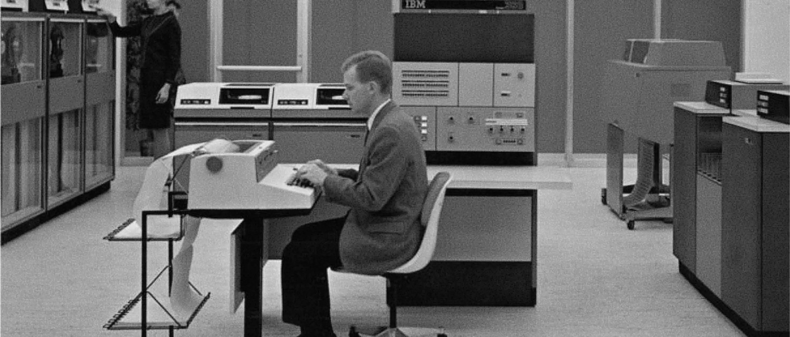 An archival photograph of a person working at a computer in a machine room.